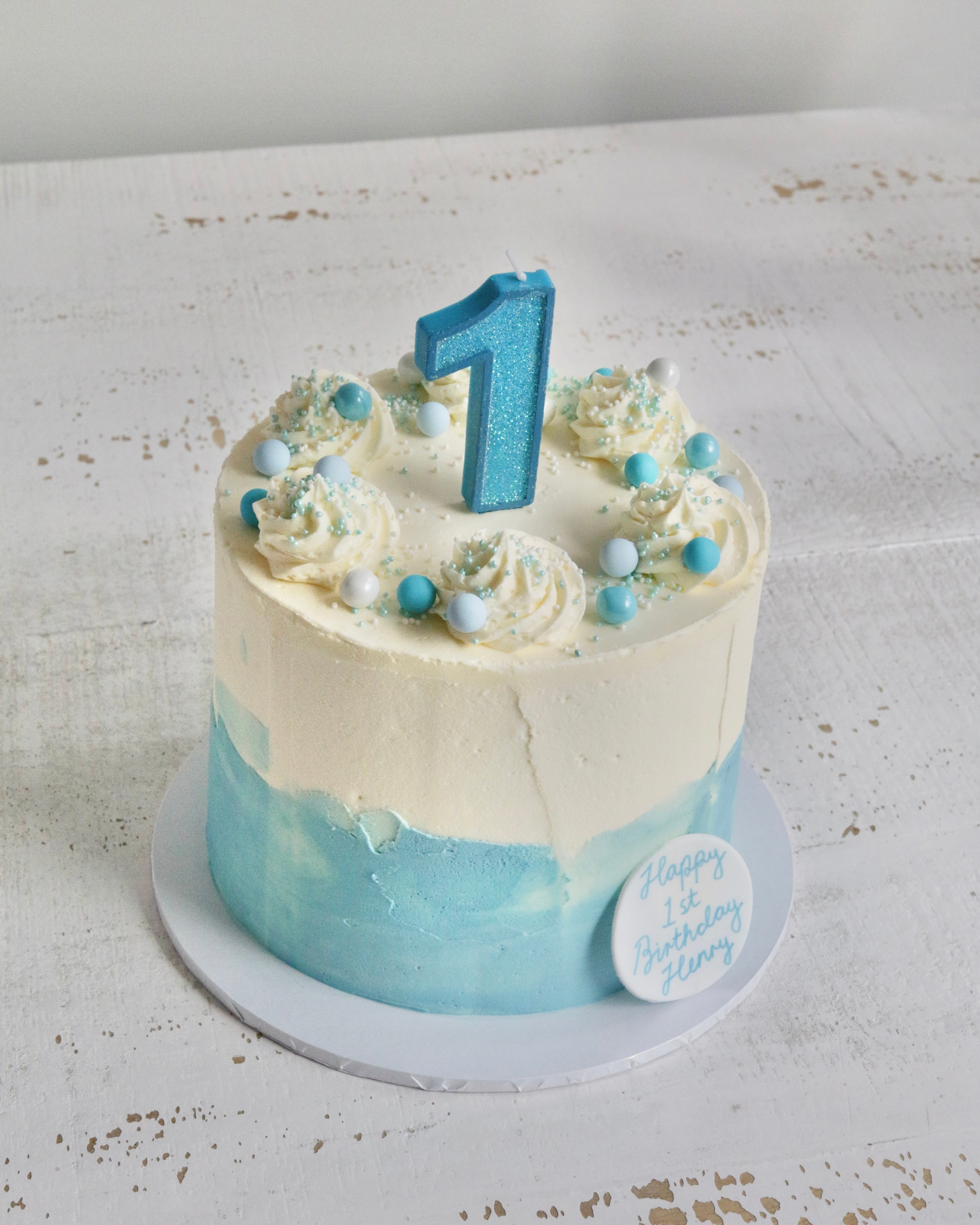 Blue Buttercream 1st Birthday Cake. A round birthday cake with white frosting on top and a light blue watercolour-style lower half, decorated with piped swirls, small blue and white beads, and a glittery blue number 1 candle. A round white sticker on the side reads 'Happy 1st Birthday Henry'.