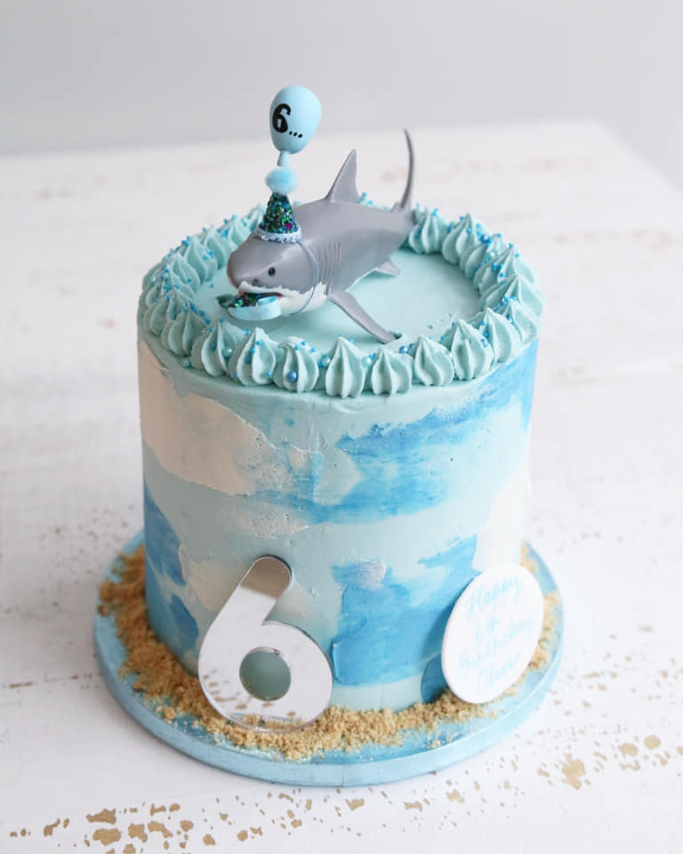 Blue Buttercream Cake with a Toy Shark on top in a party hat holding a blue balloon with a number 6.