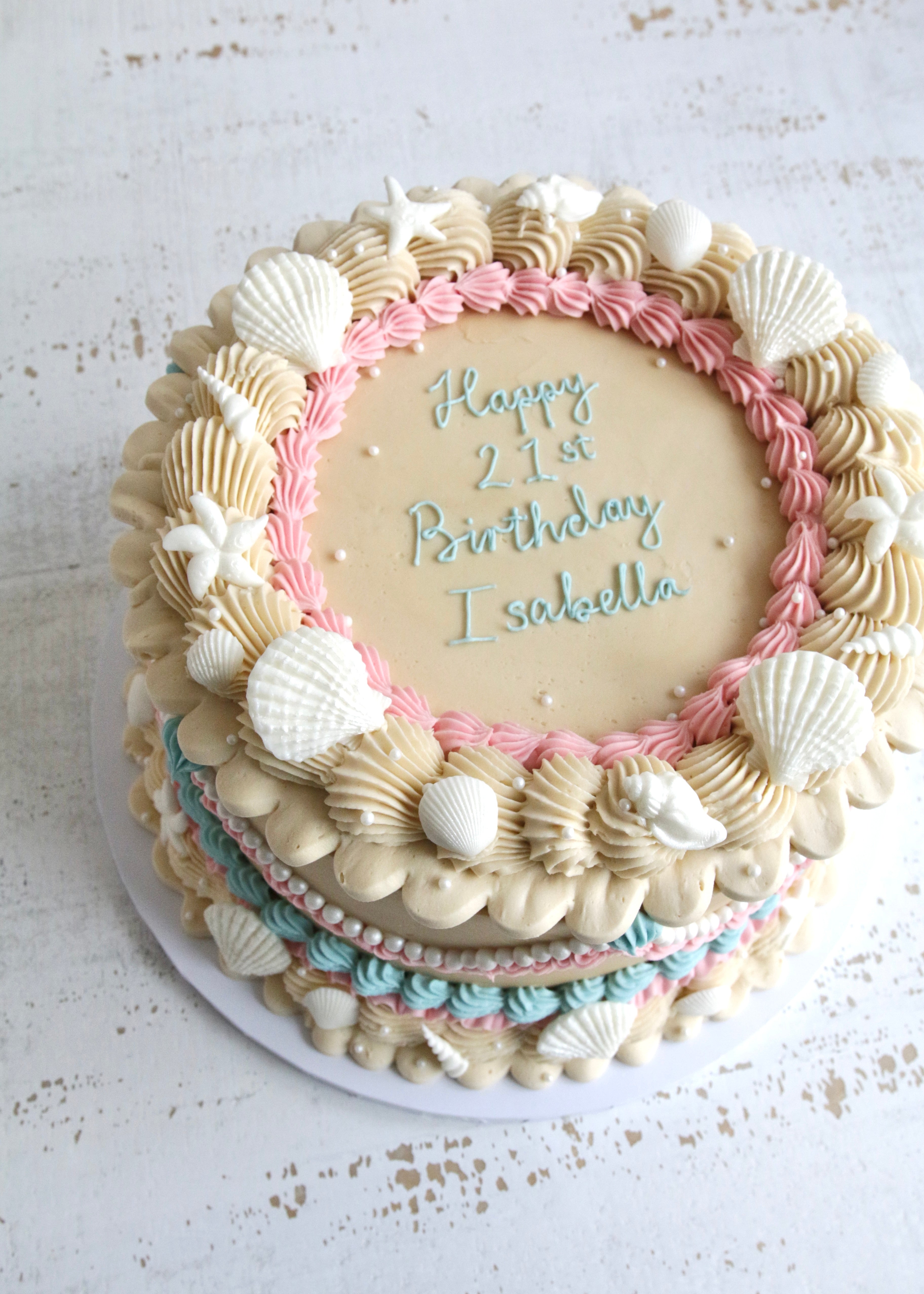 Buttercream Shell Cake with Mermaid Vintage Piping. A round birthday cake decorated with sand coloured frosting, pink and blue piped borders, white seashells, starfish, and blue script reading 'Happy 21st Birthday Isabella'.