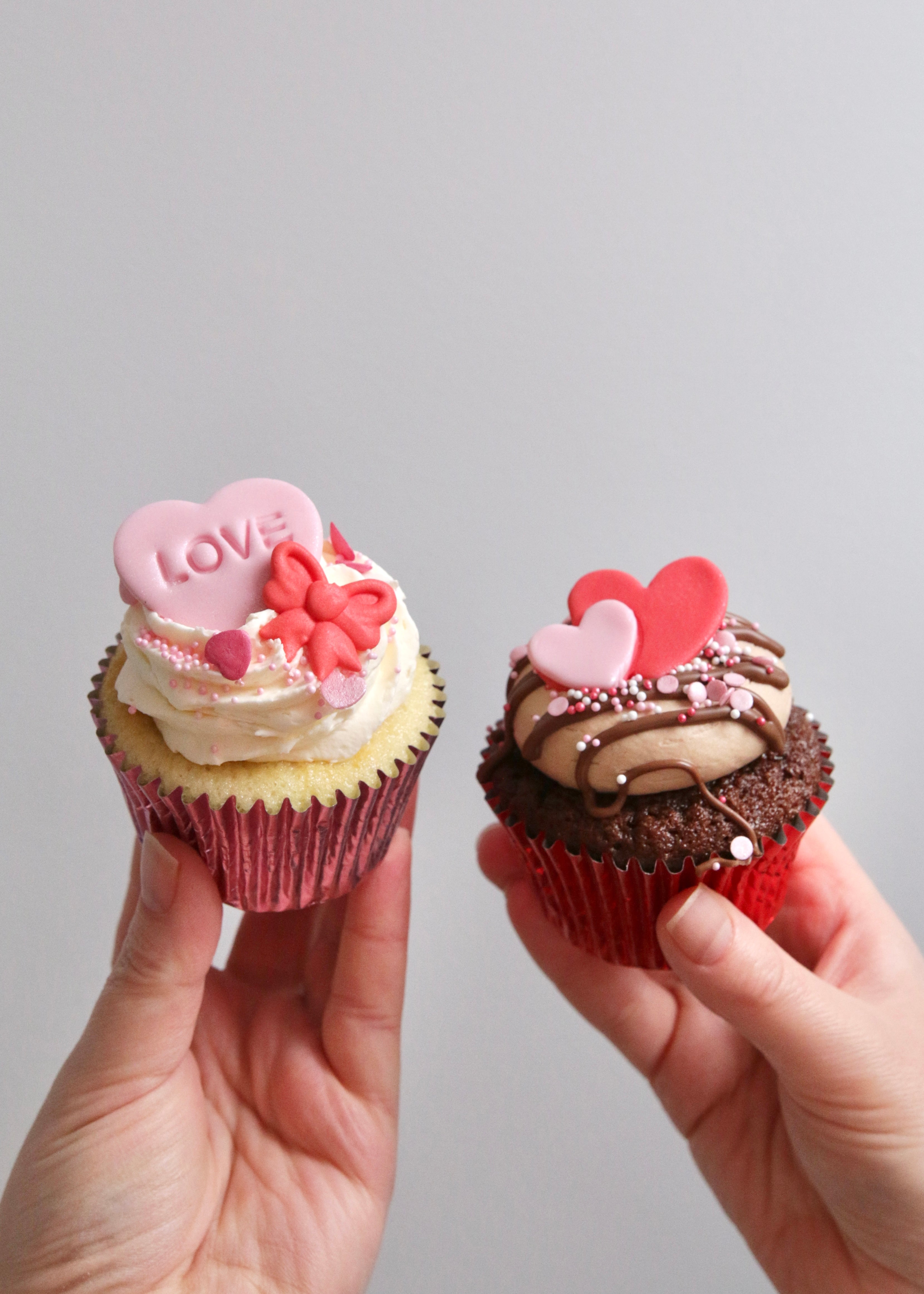 Hands Holding Two Valentine's Cupcakes - Vanilla with Pink LOVE Heart & Red Bow Decoration and Chocolate with Pink & Red Heart Decoration