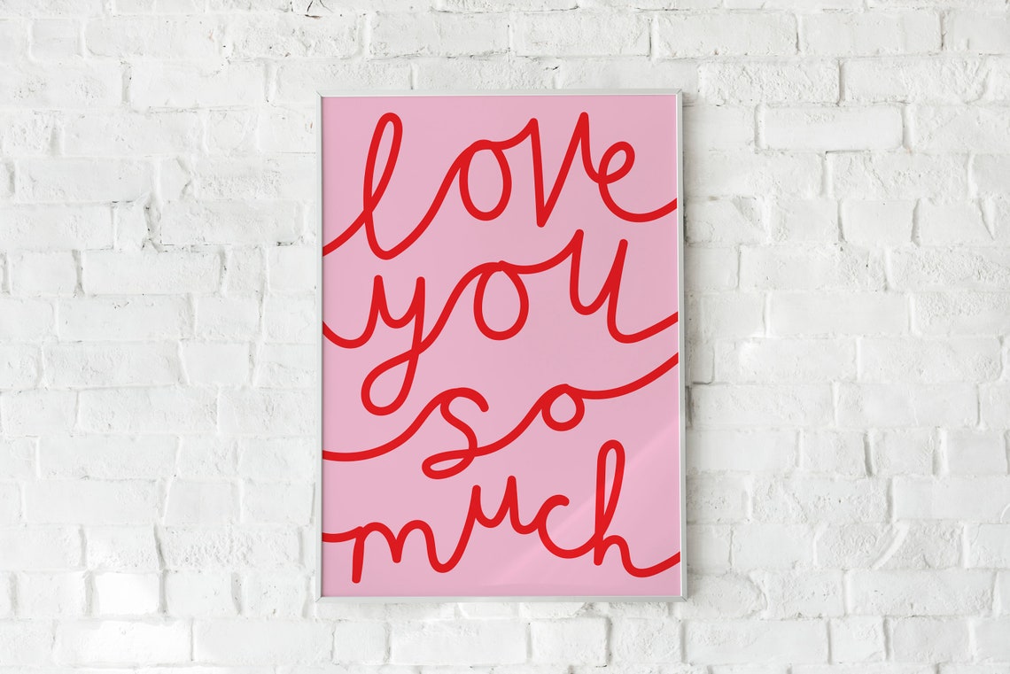 A vertical art print with the phrase 'love you so much' in red cursive script on a light pink background, displayed against a white brick wall.
