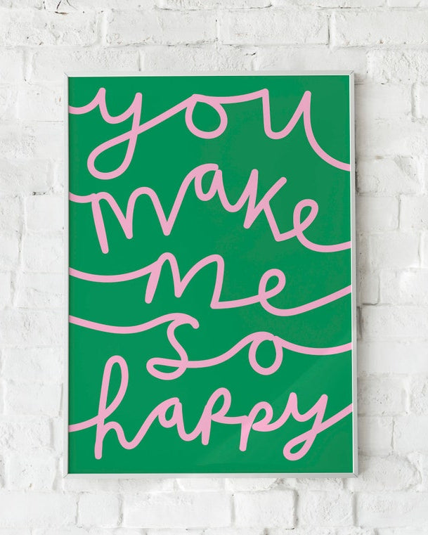 A vertical art print with the phrase 'you make me so happy' in pink cursive script over a solid green background, displayed on a white brick wall.