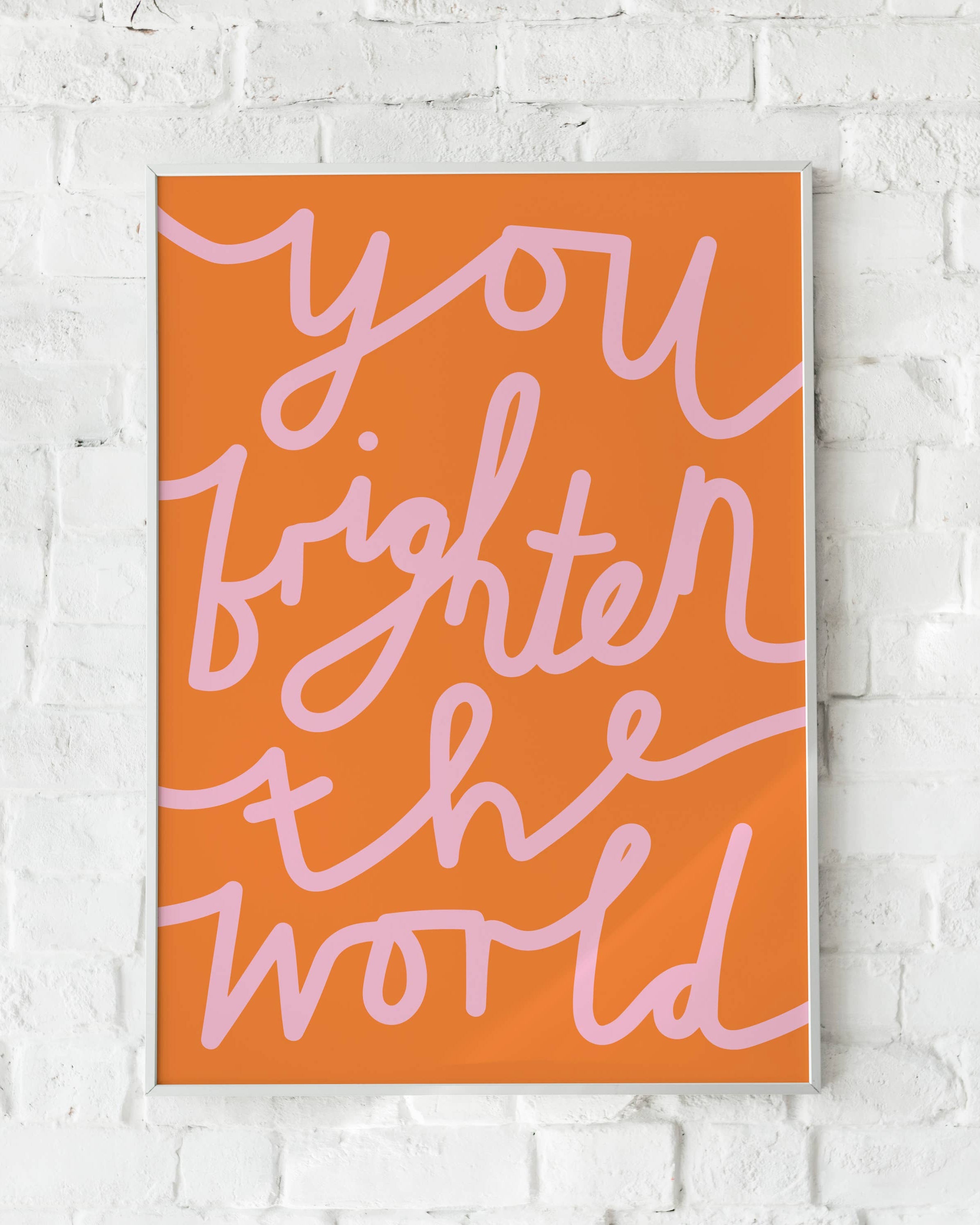 A framed art print featuring the quote 'you brighten the world' in light pink script on a vibrant orange background, displayed against a white brick wall by designer Lottie Hall