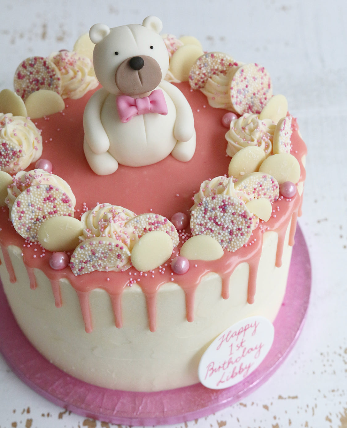 1st Birthday Vanilla Cake with a Pink Drip Decorated with White Chocolates and a White Teddy in a Pink Bow Tie